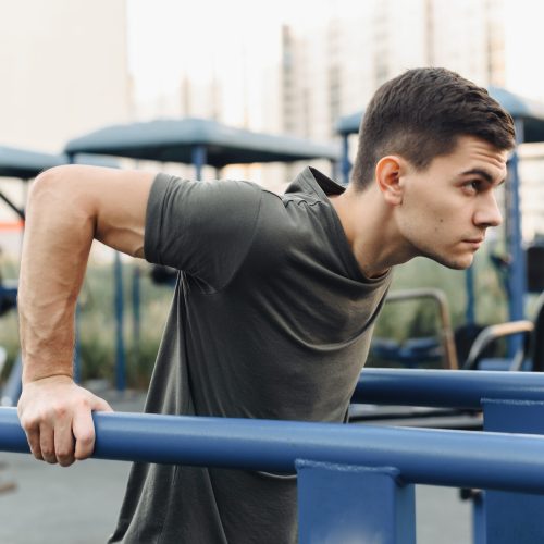 Handsome young man doing dips on the uneven bars on the outdoor sports field. Healthy lifestyle, sports and endurance concept.